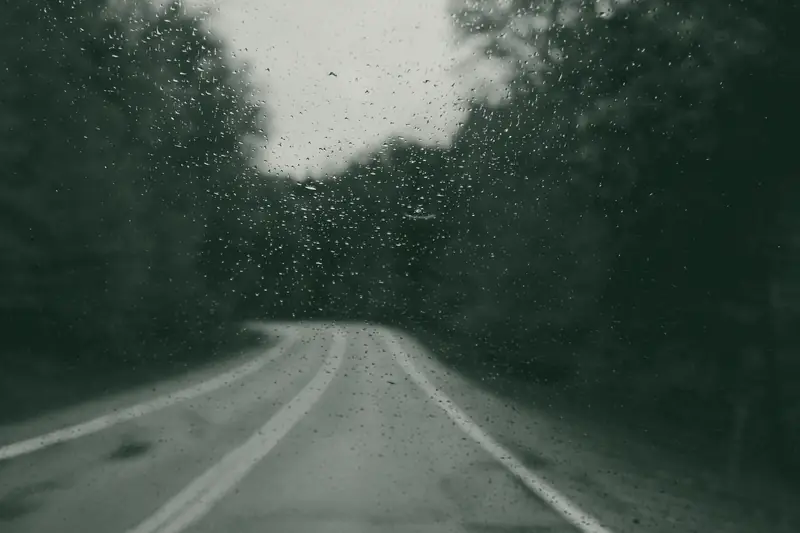 A two-lane road through autumn woods, seen through a rain-speckled windshield